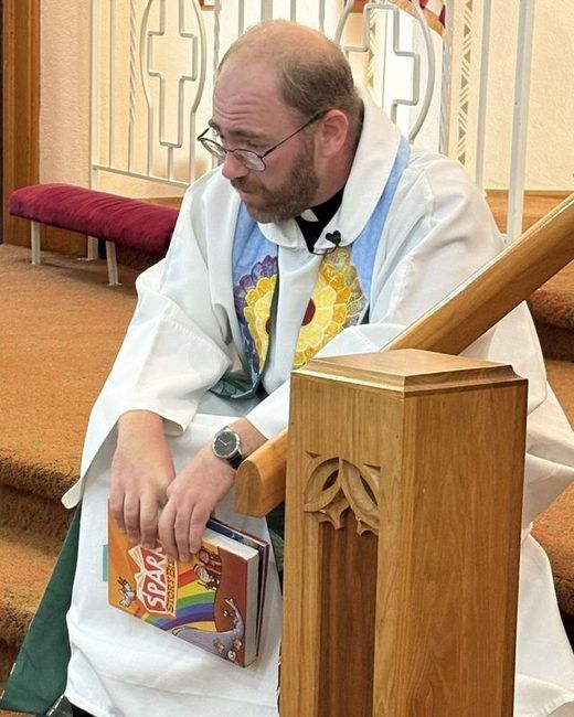 Pastor Will in his alb holding a hymnal beside the chancel rail