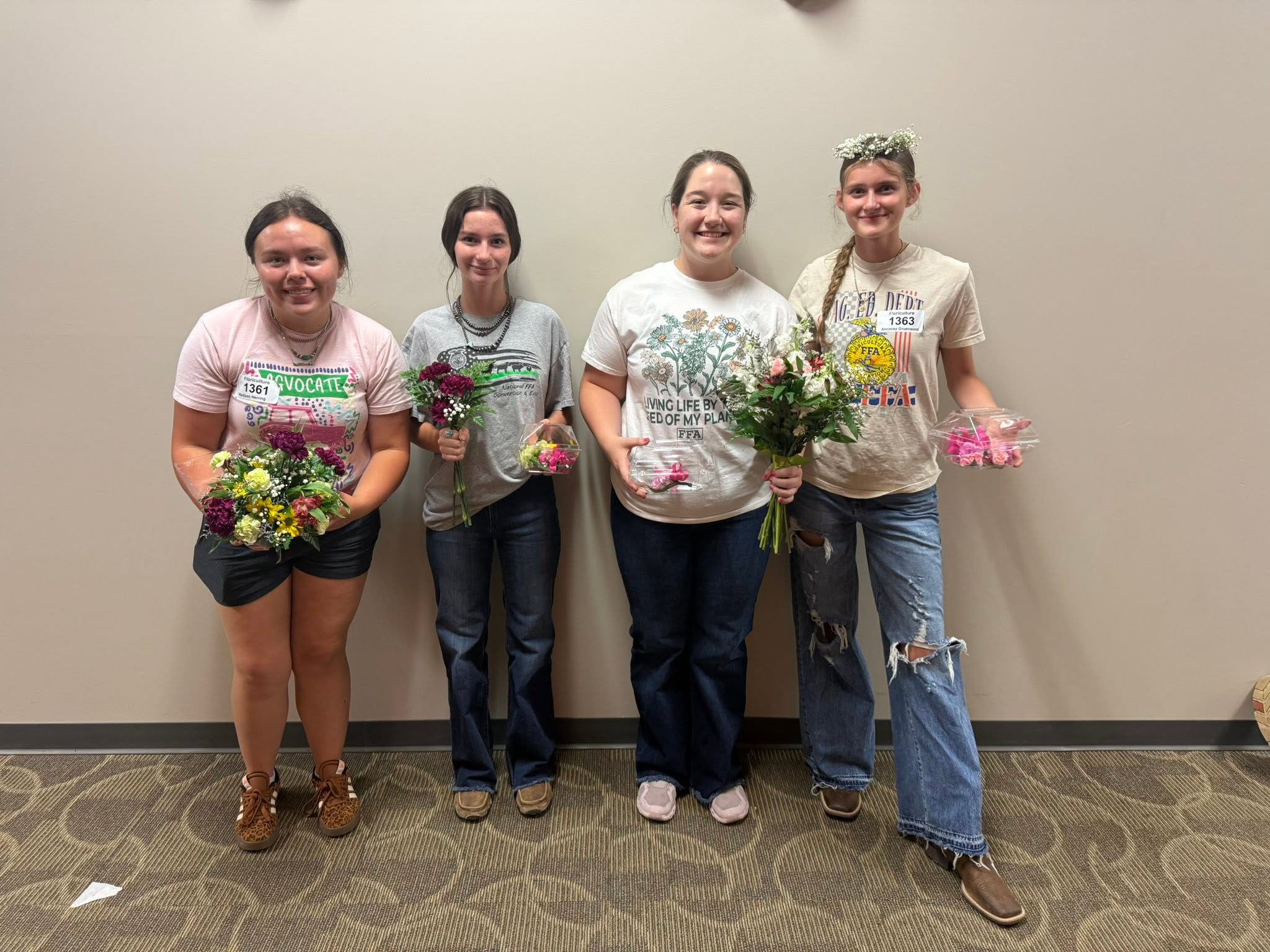 Four members holding small bouquets against a plain wall.