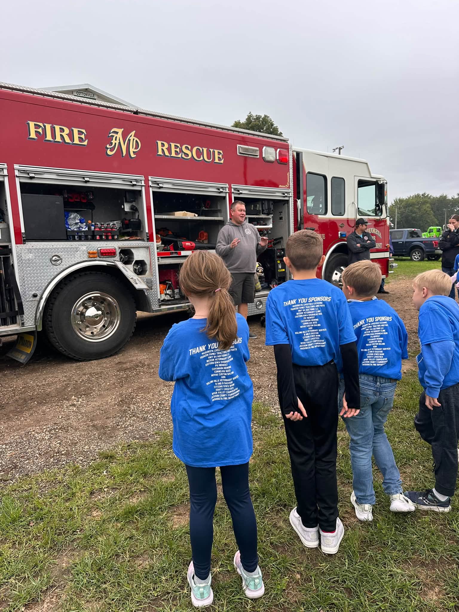 Children in blue shirts stand facing a red Maquoketa Fire Department rescue truck.