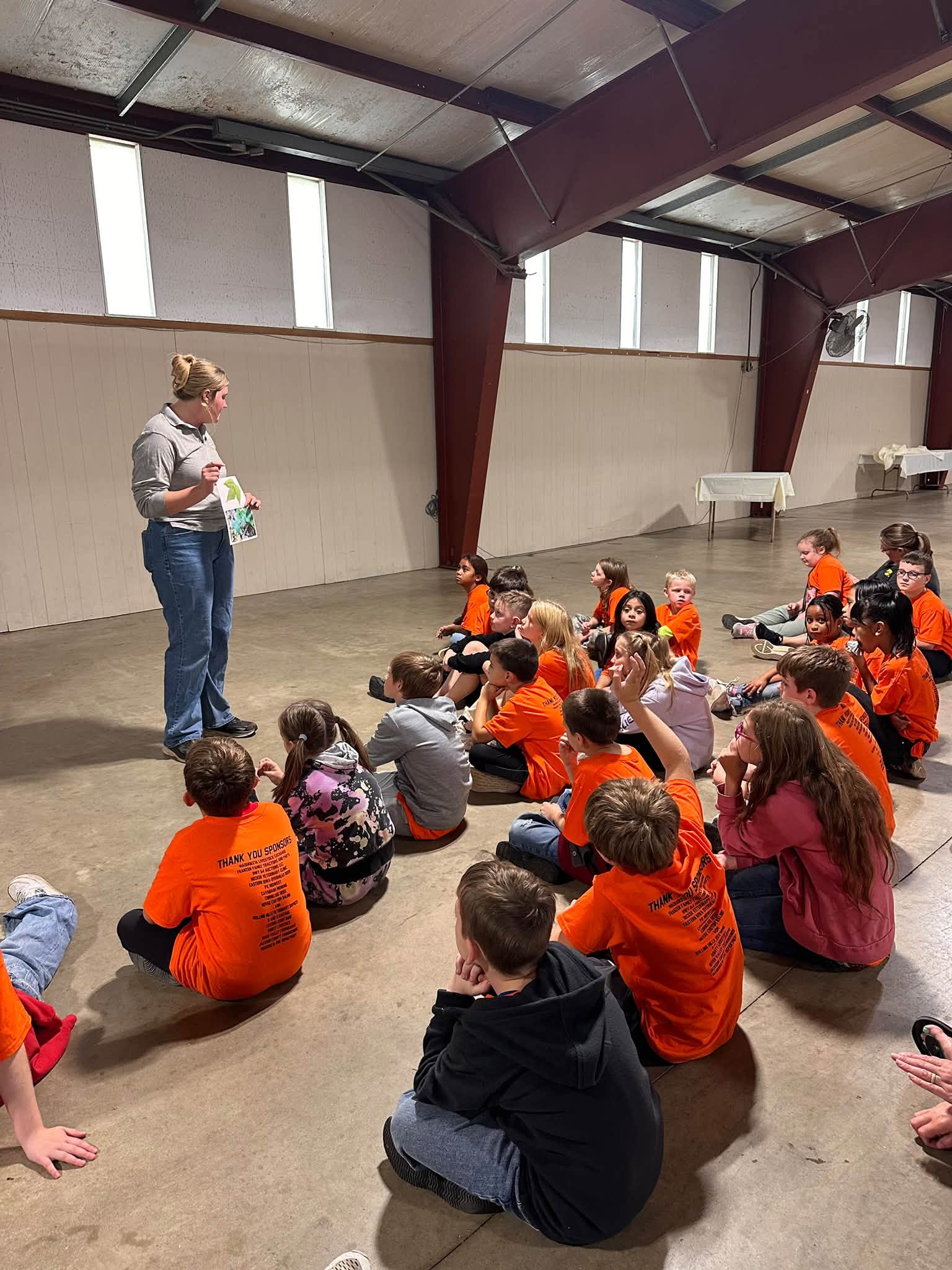 Children in orange shirts sit on the floor of a large hall listening to a presenter.