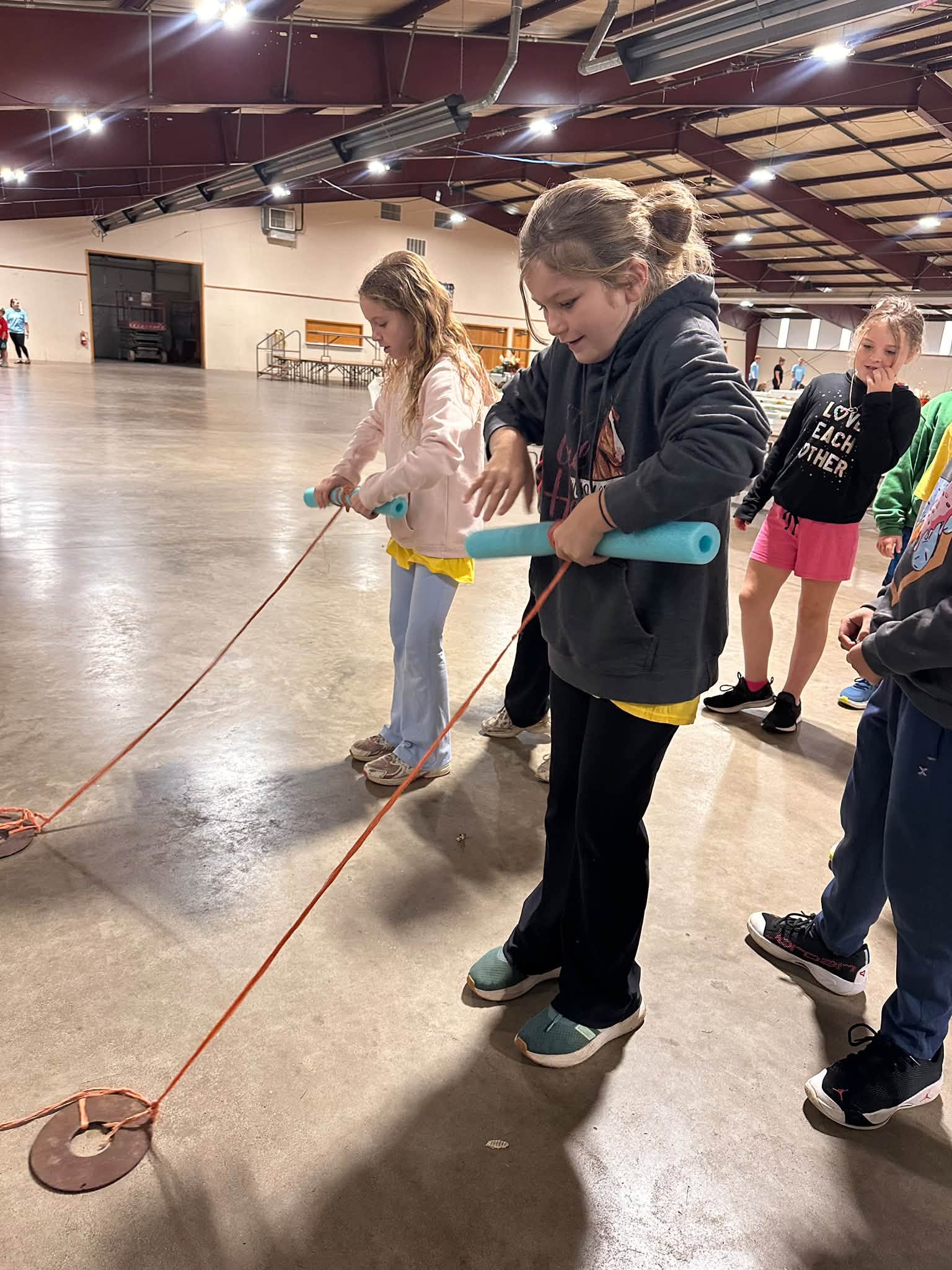 Children and volunteers handle ropes at a station in a large exhibition hall.