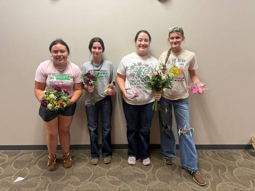 Four members holding small bouquets against a plain wall.