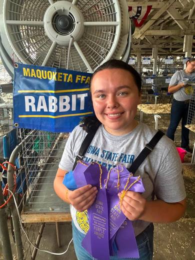 A member holding purple ribbons beside a blue Maquoketa FFA rabbit sign.