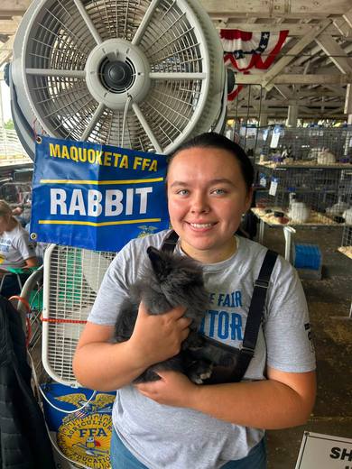 A member holding a gray rabbit beside a Maquoketa FFA rabbit sign.