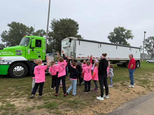 Children in pink shirts gather beside a green semi truck and grain trailer.