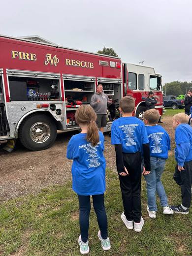 Children in blue shirts stand facing a red Maquoketa Fire Department rescue truck.