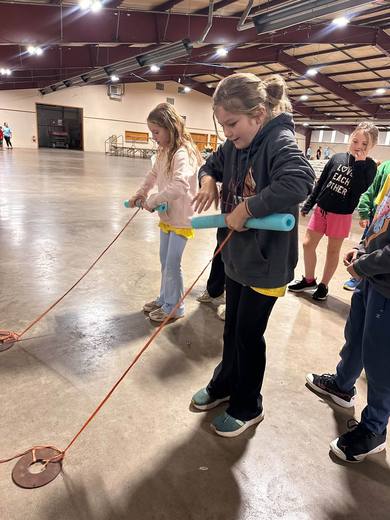 Children and volunteers handle ropes at a station in a large exhibition hall.