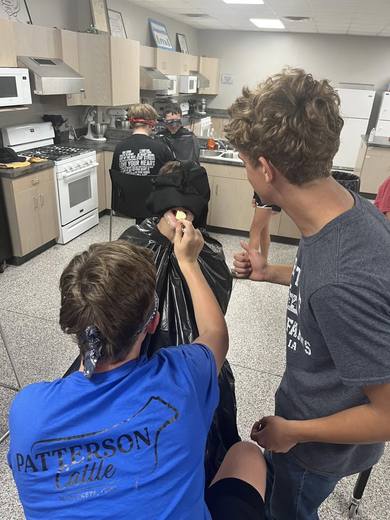 Students in a classroom kitchen taking part in a game, one wearing a blue shirt.